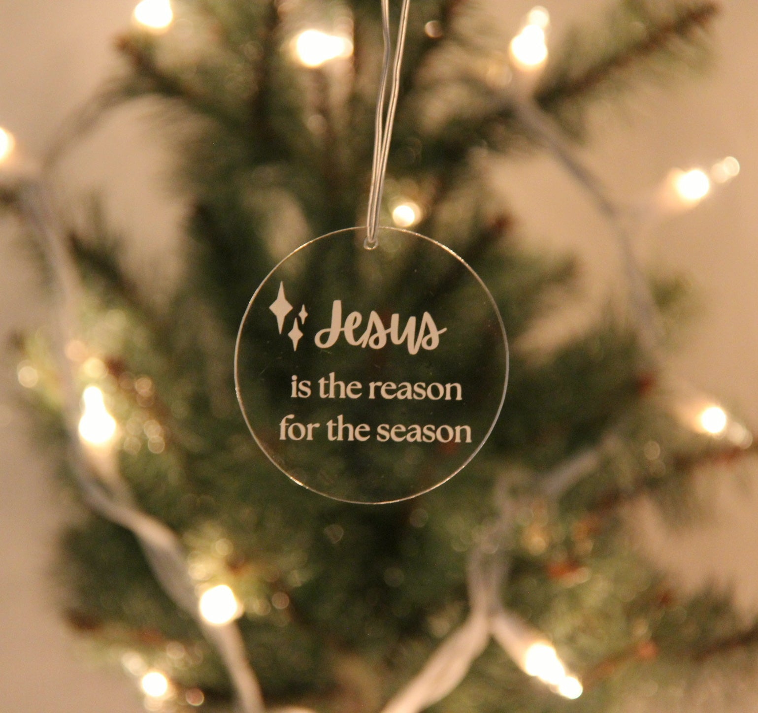 Decorative Christmas ornament with 'Jesus is the reason for the season' text on a blurred Christmas tree background.