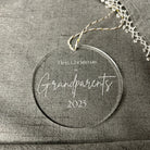 Round ornament with 'First Christmas as Grandparents 2023' text on a gray fabric background
